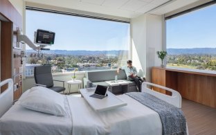 A patient room with bed, equipment, and large glass windows bringing in light