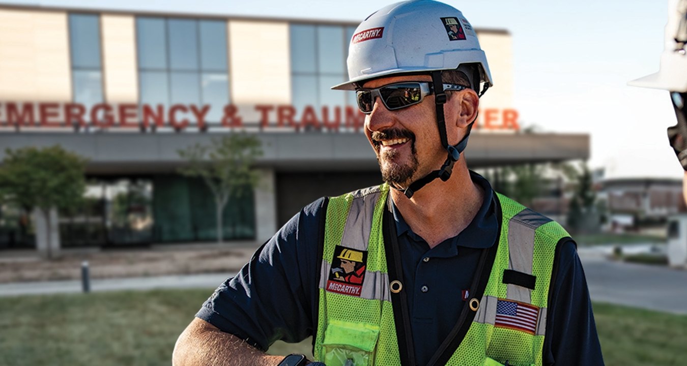 Two people in construction PPE standing outside in front of an emergency and trauma center. 