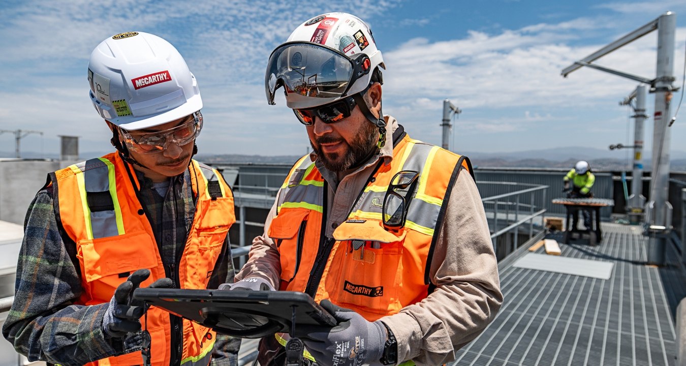 Two people wearing construction PPE at a jobsite while looking at a tablet.