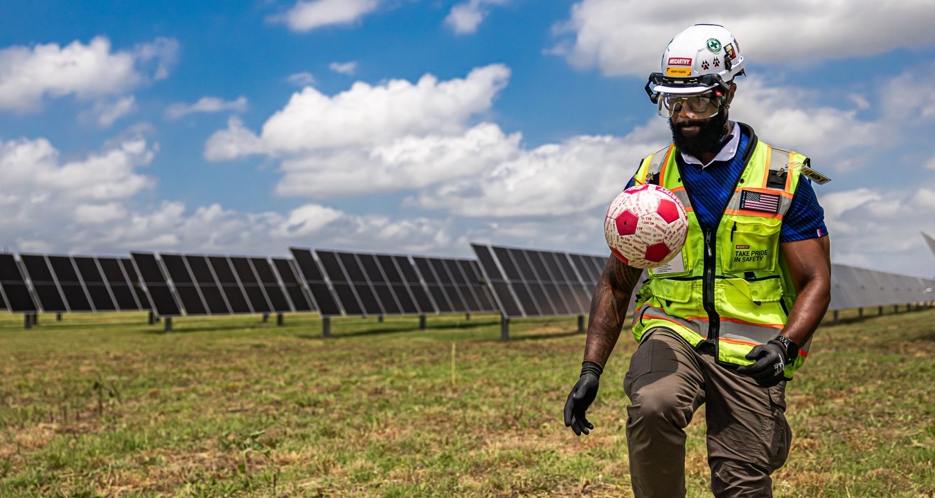 A person wearing PPE and kicking a soccer ball in front of a solar field.