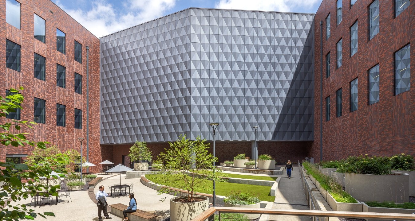 Exterior courtyard between brick office buildings, showing a faceted metal-clad corner facade above a landscaped plaza with people seated and walking.