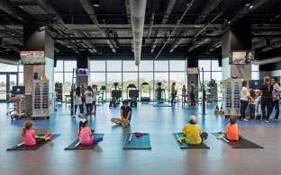 An open exercise room with people sitting on yoga mats and exercise equipment in the background