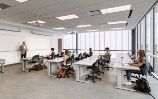 People in a classroom with desks and chairs, and a teaching presenting in front of the class