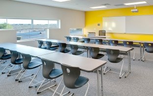 Classroom with rows of tables and chairs and a white board with a yellow wall behind it