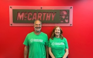 Two people wearing Construction Inclusion Week shirts, standing in front of a McCarthy sign.