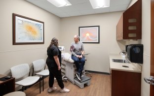 A person sitting on the bed of a patient room and another person standing next to them.