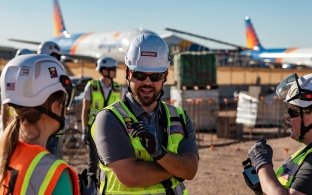 Three people having a discussion at an airport jobsite with planes in the background