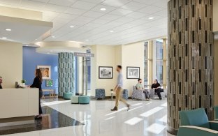 A waiting area and check in desk with someone talking to the receptionist and other people sitting in the chairs