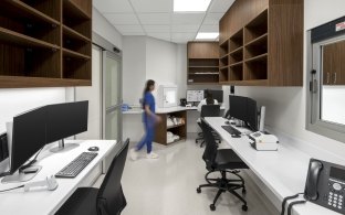 A modern healthcare office area featuring neutral tones with natural wood elements. The space includes desks with computers. Multiple staff members are seen.