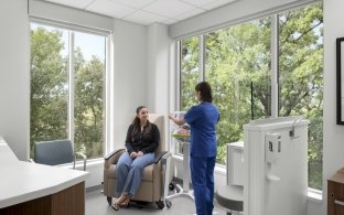 A modern healthcare infusion room featuring expansive glass windows and green trees outside. A nurse is assisting a patient.