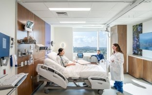 Bright patient room with nurse talking to a patient in bed and a visitor seated by a large window.