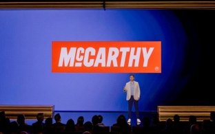 A person presenting on stage with the McCarthy logo in the background.