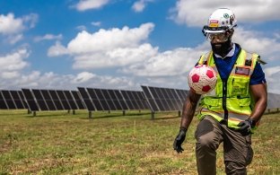 A person wearing PPE and kicking a soccer ball in front of a solar field.