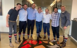 	Seven McCarthy colleagues stand together in an office hallway, all wearing matching airplane-print socks.