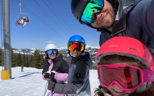 A family of four in ski helmets and goggles smiles for a photo on the slopes, with a chairlift in the background.