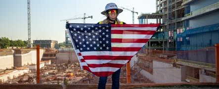 A person wearing PPE and holding a U.S. Flag in front of them with a jobsite in the background.