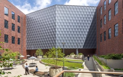 Exterior courtyard between brick office buildings, showing a faceted metal-clad corner facade above a landscaped plaza with people seated and walking.