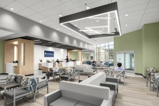 A modern healthcare waiting room featuring neutral tones with green accent walls and natural wood elements. The space includes rows of cushioned chairs with geometric-patterned upholstery, a sleek reception desk with multiple staff members assisting patients, and a large, angular overhead light fixture. Floor-to-ceiling windows bring in natural light, and the signage on the wall reads “The University of Kansas Health System.”