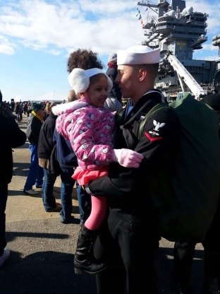 A person in a military uniform holding a young child with a ship in the background