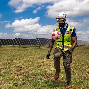 A person wearing PPE and kicking a soccer ball in front of a solar field.