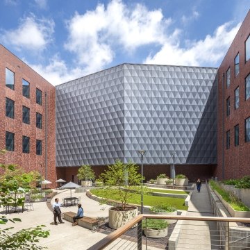 Exterior courtyard between brick office buildings, showing a faceted metal-clad corner facade above a landscaped plaza with people seated and walking.